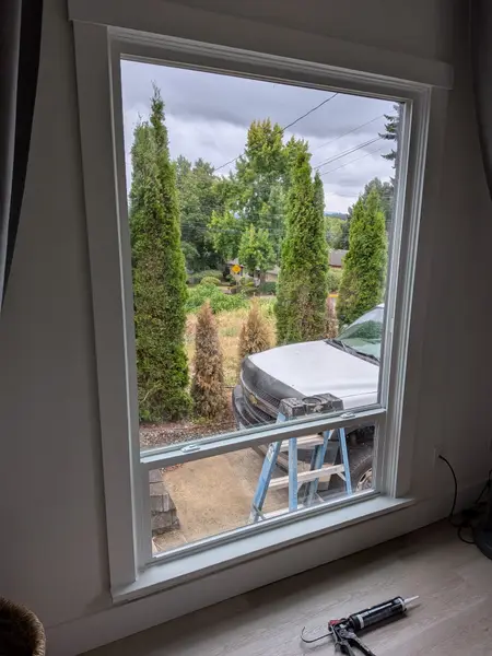 Caulk gun resting on the sill of a newly set window, seen from inside looking out at the job truck and ladder