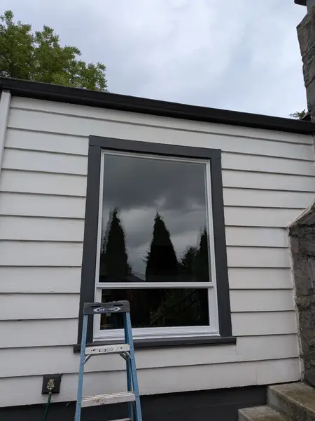 Finished window with dark charcoal trim on white lap siding, ladder still standing after the install