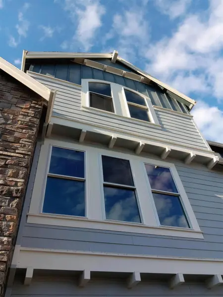 Looking up at a gray lap-siding house with stone veneer: three white double-hung windows below a pair with an arched top, under a blue sky
